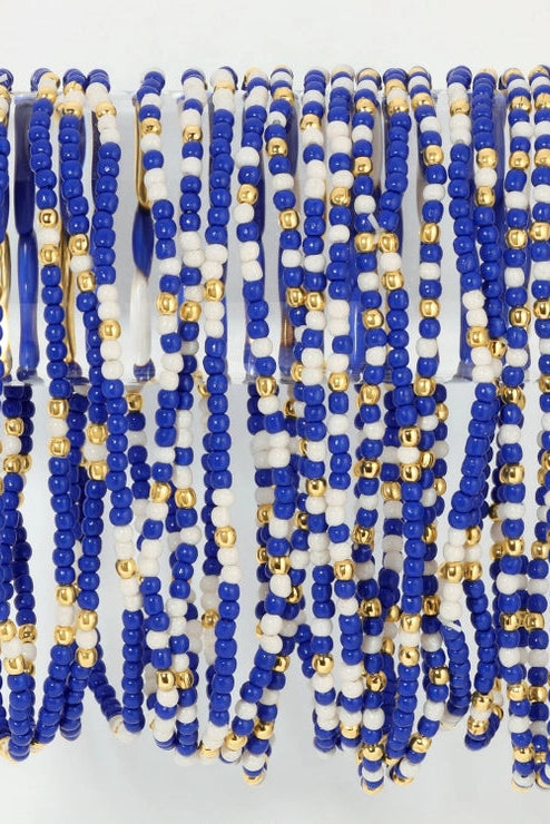Multiple strands of beaded bracelets draped over a clear acrylic bar. The bracelets feature alternating rows of royal blue and white round beads, with small metallic gold spacer beads throughout.