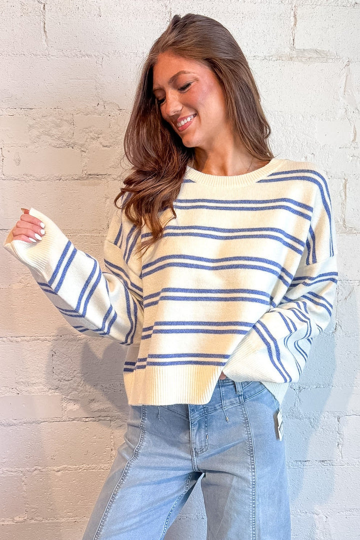 A woman wearing a cream and blue striped long-sleeve knit sweater and light-wash wide-leg jeans, posing against a white brick wall.