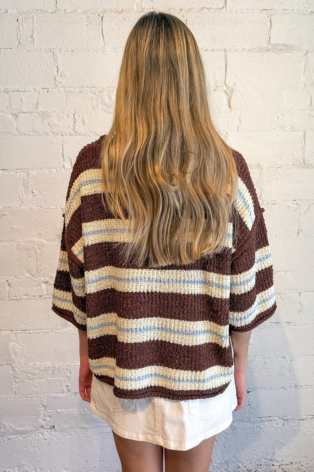 A woman viewed from behind wearing a brown, cream, and light blue striped knit cardigan sweater, standing against a white brick wall.