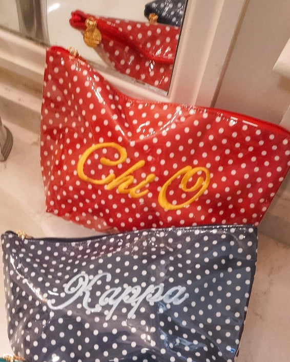 Three personalized cosmetic pouches with white polka dots are lined up on a bathroom sink. The top red pouch is embroidered with 'Chi Omega' in yellow, the middle navy blue pouch is embroidered with 'Kappa' in white, and the bottom teal blue pouch is embroidered with 'Zeta' in white.