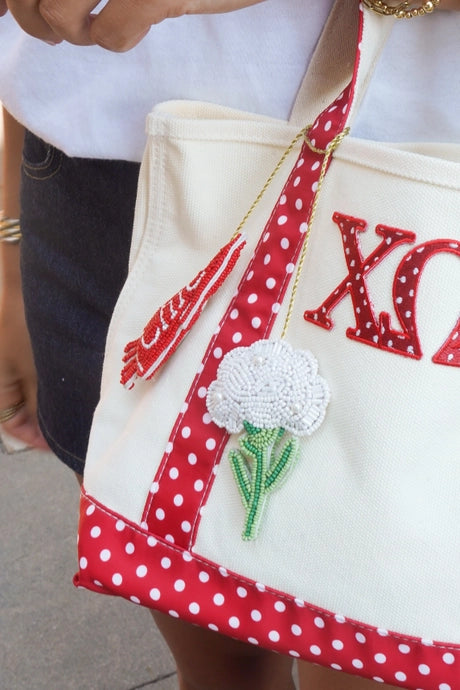 A cream canvas tote bag with red and white polka dot straps and trim, featuring a red sequined Greek letter 'XO' and a white flower applique. The bag is being held by a person wearing gold bracelets and a white shirt.