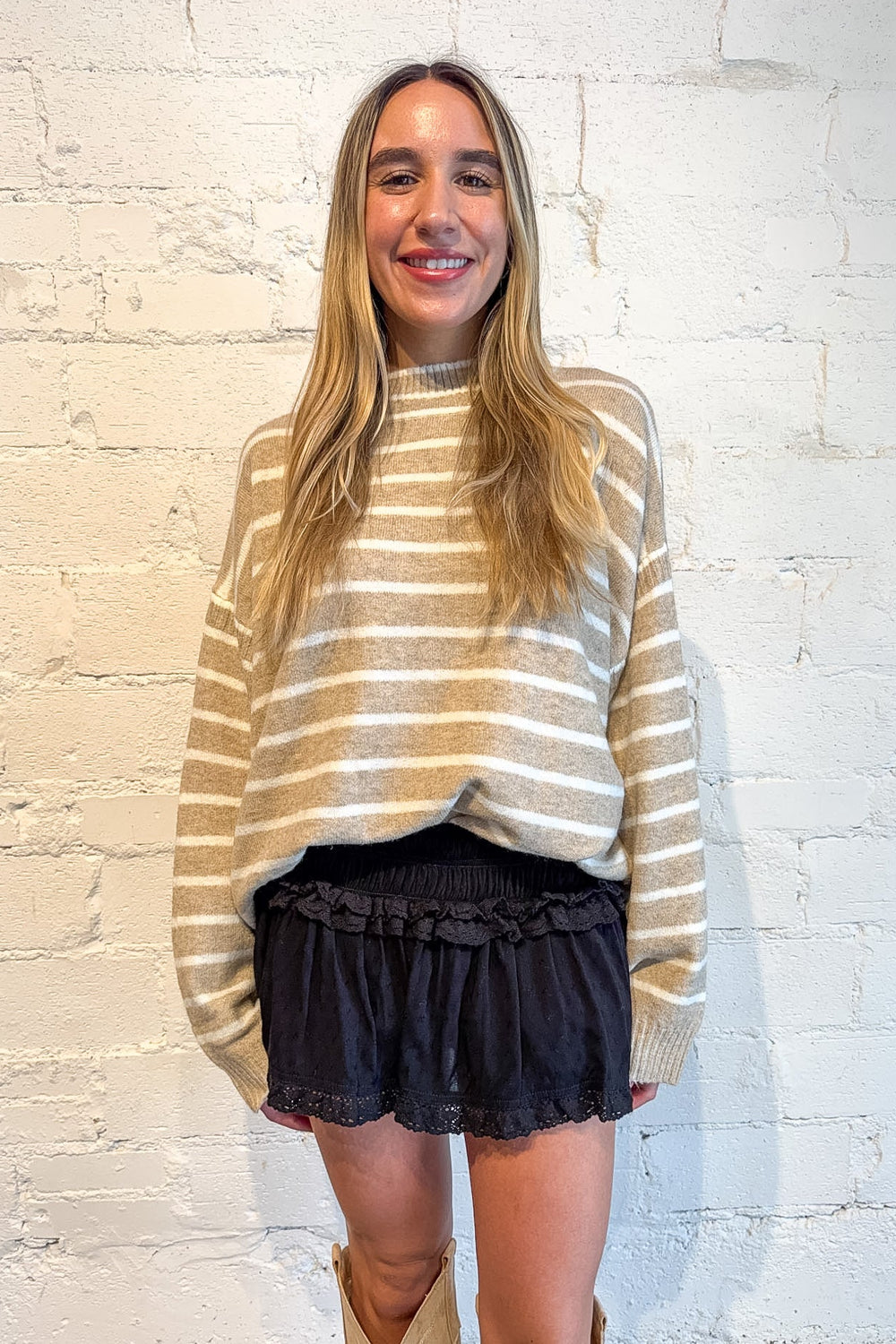 A woman stands against a white painted brick wall wearing a beige and white horizontally striped knit sweater, dark ruffled shorts, and tan cowboy boots.