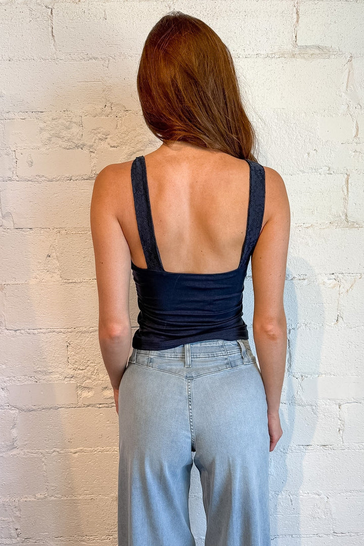 A woman viewed from behind wearing a dark navy racerback tank top and light-wash wide-leg jeans, standing against a white brick wall.