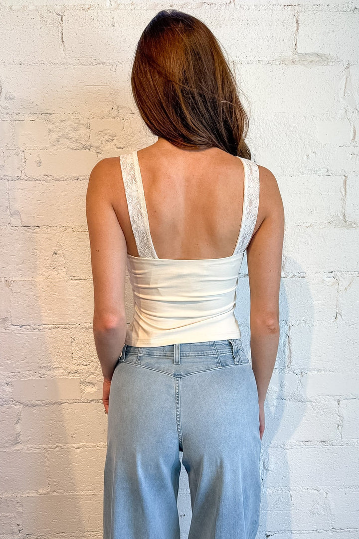 A woman shown from behind wearing a white lace-trimmed scoop-neck tank top and light-wash wide-leg jeans, standing against a white brick wall.