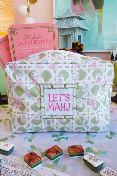 A patterned fabric mahjong case with a white, sage green, and pink lattice design, featuring a central patch that reads 'LET'S MAHJ'. Several mahjong tiles are arranged in front of the case on a patterned surface, with a pink book titled 'let's do the CHARLESTON' visible behind it.