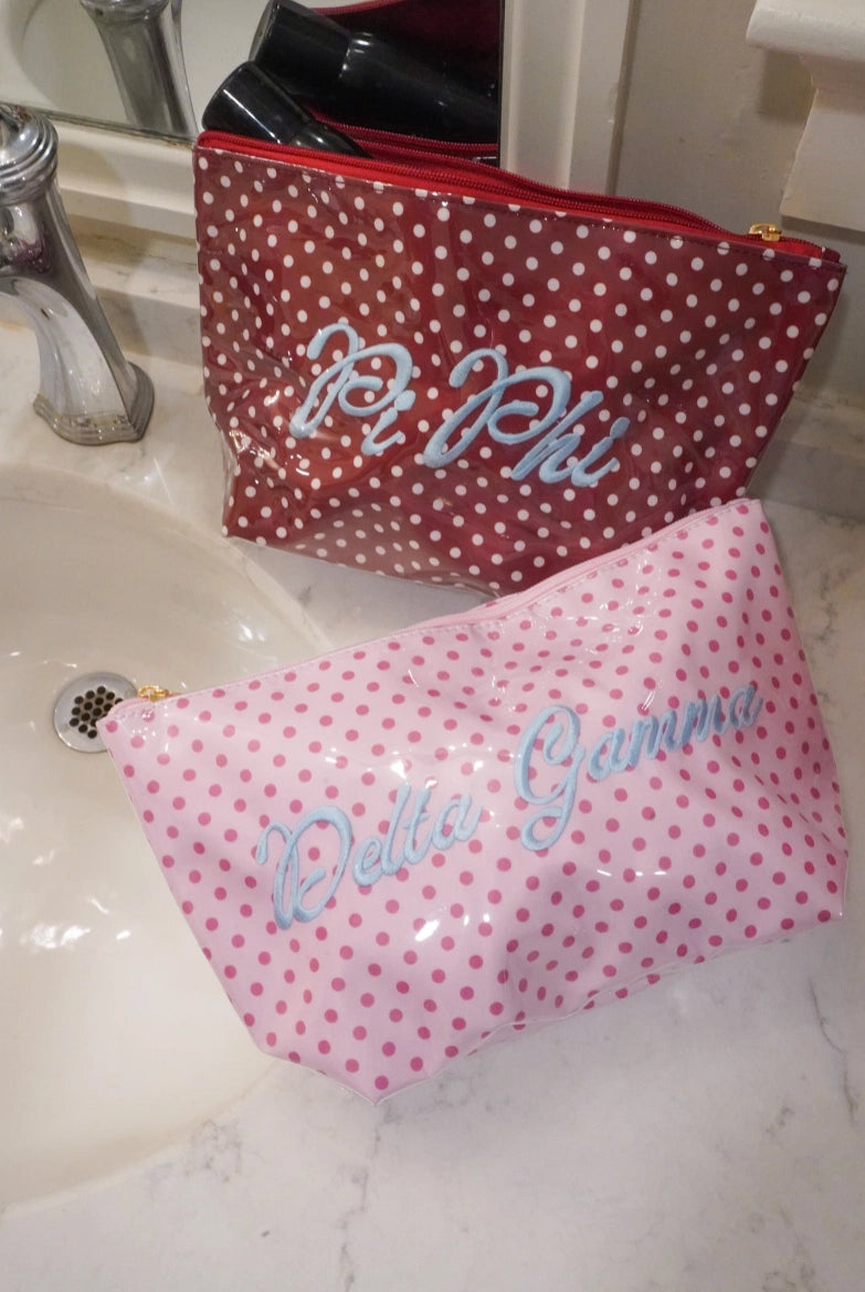 Two polka dot cosmetic pouches on a bathroom sink countertop. The back pouch is dark red with white polka dots and silver script reading 'Pi Phi'.