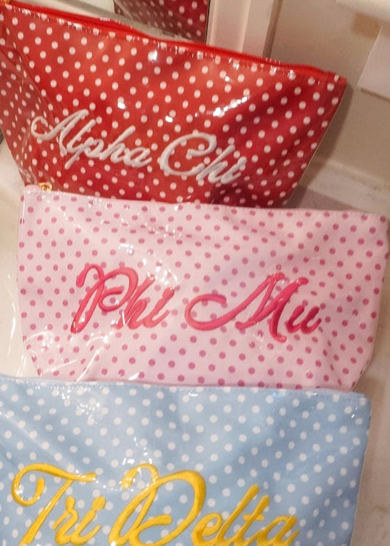 Three personalized zippered pouches with white polka dots are stacked on a bathroom sink. The top red pouch is labeled 'Alpha Chi' in white script, the middle pink pouch is labeled 'Phi Mu' in pink script, and the bottom blue pouch is labeled 'Tri Delta' in yellow script.