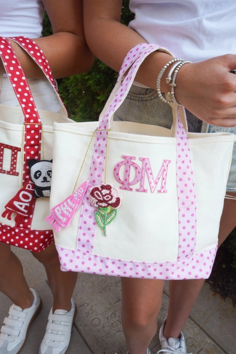 A cream canvas tote bag with pink polka-dot handles and trim, featuring pink monogrammed letters and a red rose patch, held by a person outdoors.