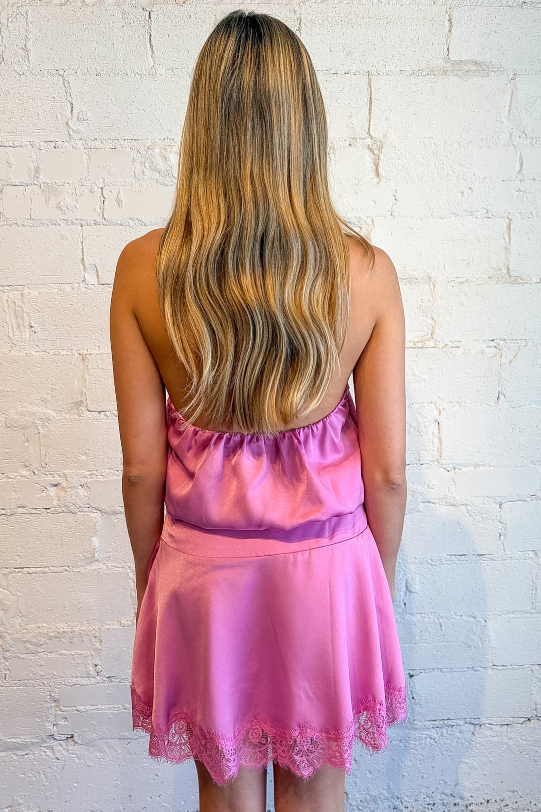 A woman with long blonde-highlighted hair is shown from behind wearing a pink satin slip dress with a lace hem, standing against a white painted brick wall.