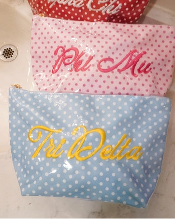 Three personalized zippered pouches with white polka dots are stacked on a bathroom sink. The top red pouch is labeled 'Alpha Chi' in white script, the middle pink pouch is labeled 'Phi Mu' in pink script, and the bottom blue pouch is labeled 'Tri Delta' in yellow script.