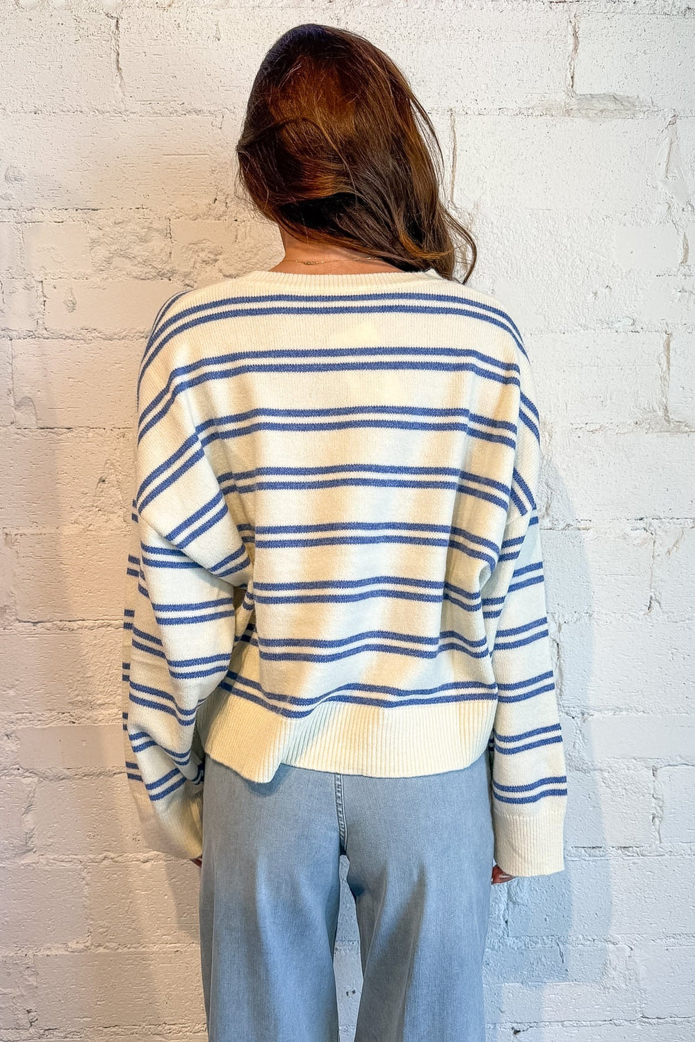 A person viewed from behind wearing a cream knit sweater with thin blue horizontal stripes and light blue wide-leg pants, standing against a white painted brick wall.