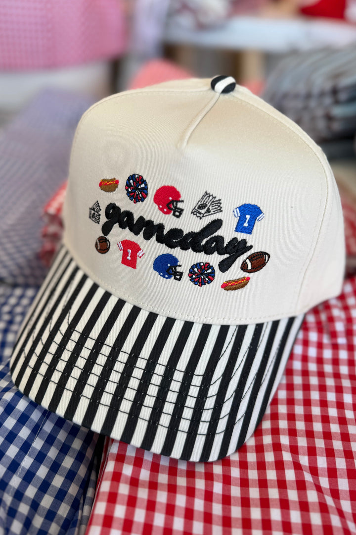 A cream-colored snapback cap with a black and white striped brim, featuring 'gameday' embroidered in black script with sports team and food icons. The cap is displayed on top of red, white, and blue gingham shirts.