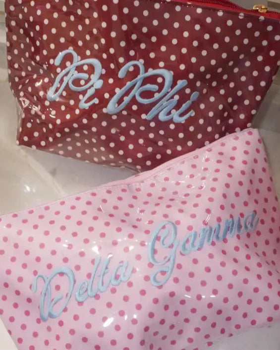 Two glossy vinyl pouches with white polka dots, one red and one pink, featuring blue cursive text reading 'Pi Phi' and 'Delta Gamma' respectively, resting on a bathroom sink.