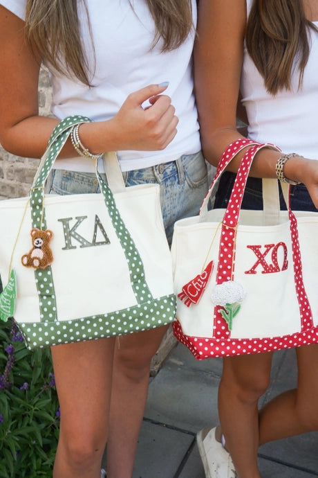 Two women holding custom canvas tote bags with green and red polka dot handles and borders. The left bag is cream with green polka dot trim and features a brown teddy bear charm with a green leaf.