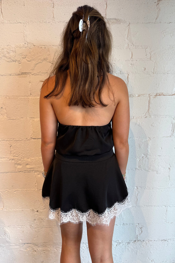 A woman viewed from behind wearing a black strapless mini dress with a white lace hem, standing against a white brick wall.